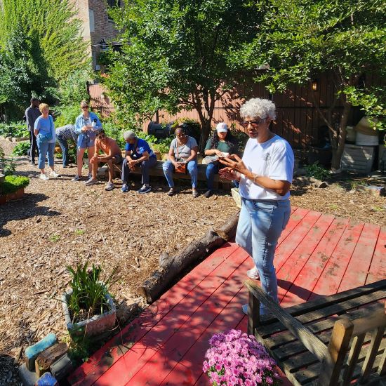 Gina Jamison, standing on a red wooden stage in a community garden, talks to a group of visitors